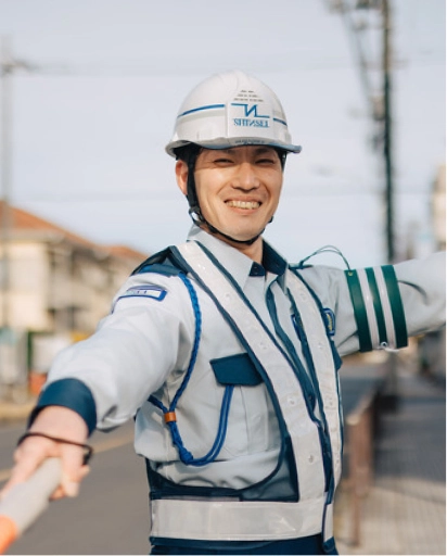 シンセイのロゴが入ったヘルメットをかぶり、笑顔で誘導する警備員の写真
